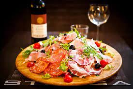 tagliere rotondo con salumi insalata e pomodorini su una tavola sullo sfondo una bottiglia di vino rosso un calice e un bicchiere